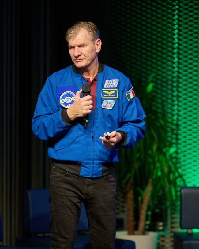 Paolo Nespoli speech aziendale con microfono in mano e giacca da astronauta su un palco illuminato di verde