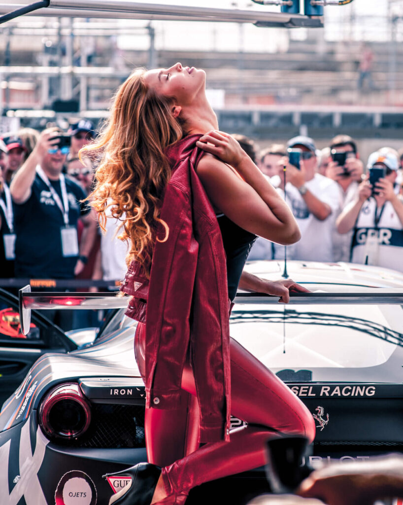 Ragazza bionda vestita Guess con pataloni di pelle e giacca rossa posa appoggiata all'alettone di una ferrari di fronte a dei paparazzi