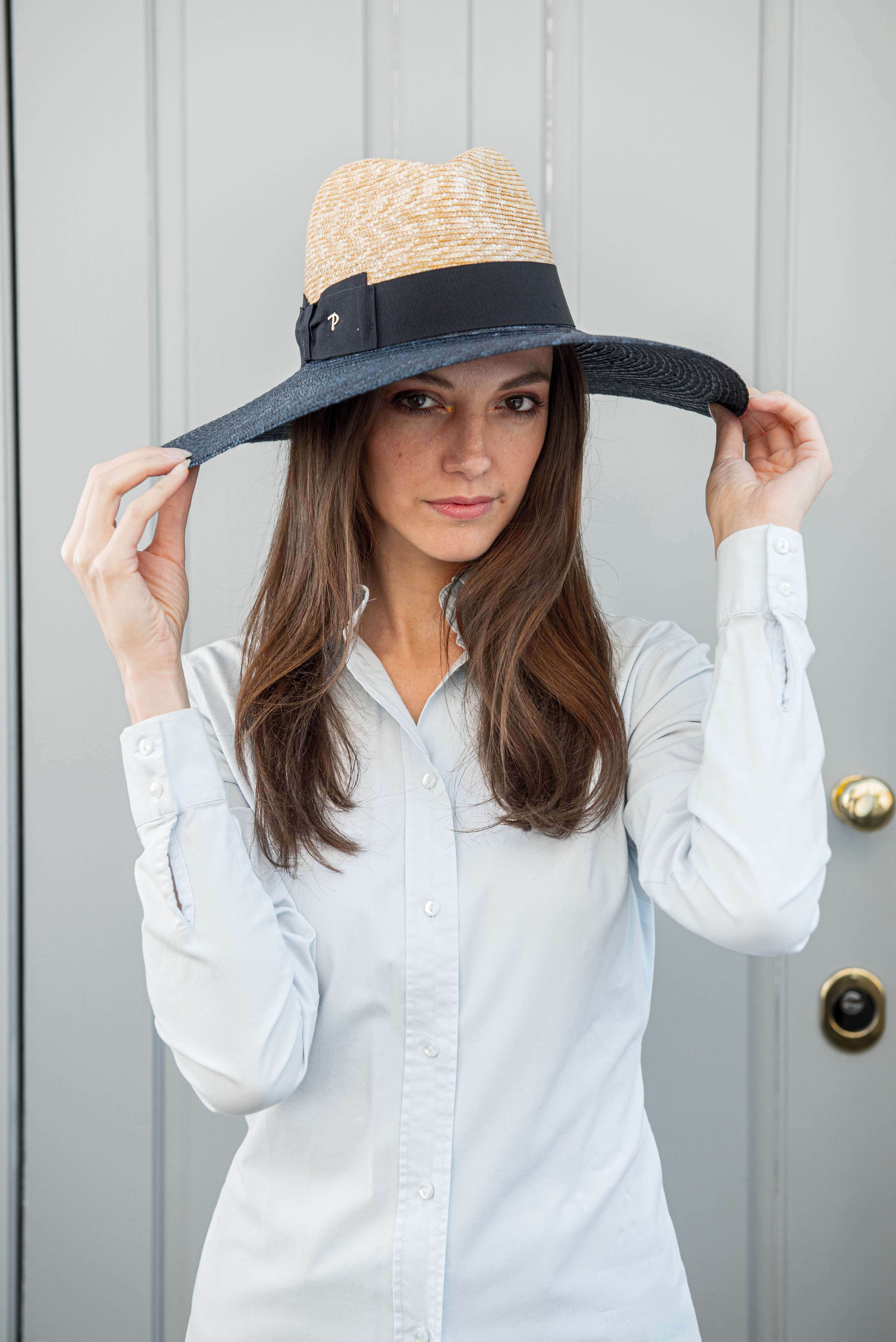 modella indossa cappello Panizza a tesa larga tenendone i lati con le mani la tesa del cappello è blu la modella indossa una camicia azzurra davanto ad un portone grigio azzurro