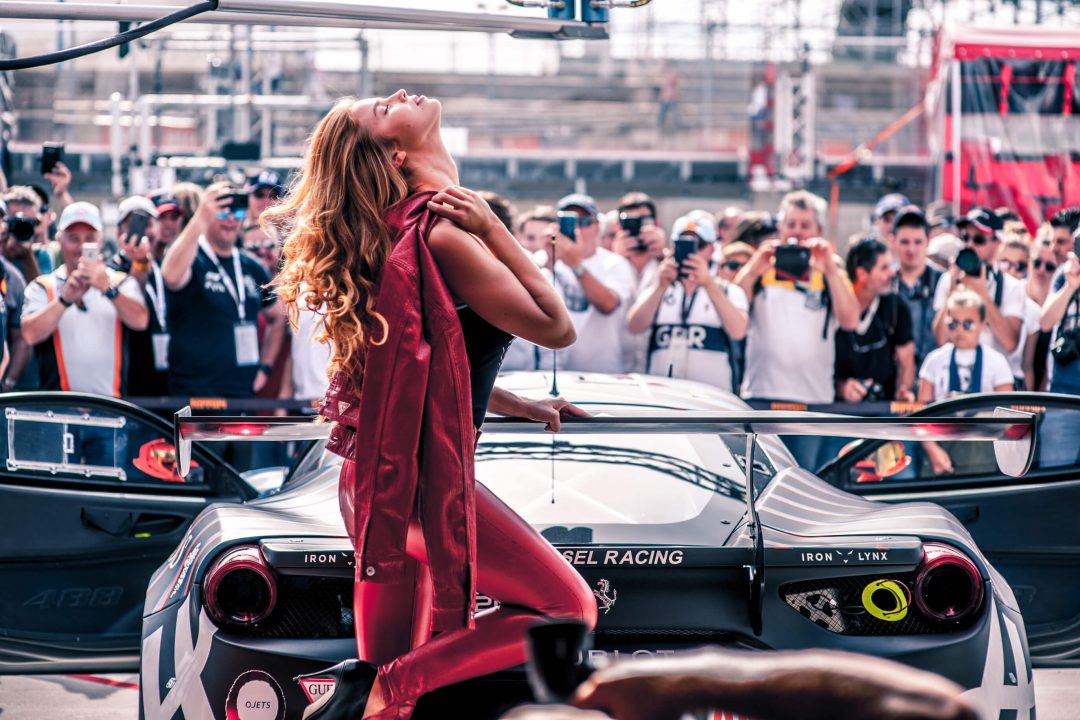 Ragazza bionda vestita Guess con pataloni di pelle e giacca rossa posa appoggiata all'alettone di una ferrari di fronte a dei paparazzi