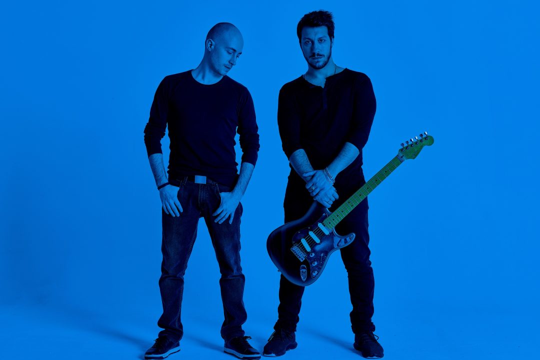 2 musicisti in piedi di fianco uno tiene la chitarra con le mani l'altro ha le mani appese alle tasche dei jeans e guarda la chitarra