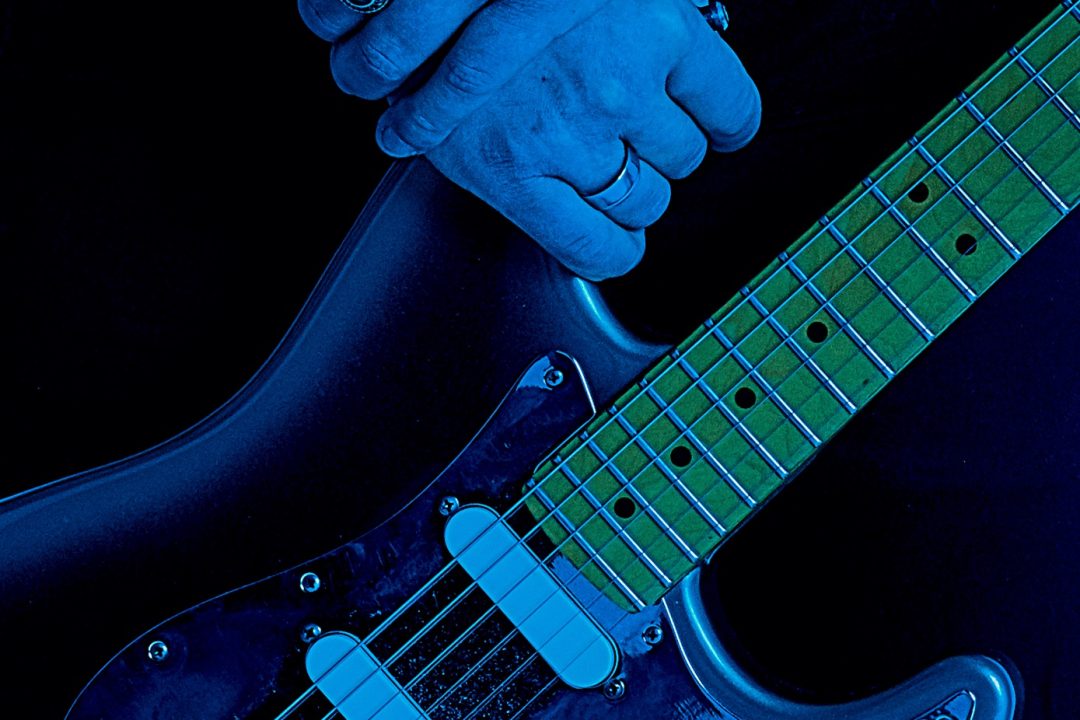 mani congiunte che tengono una chitarra elettrica la luce è blu spiccano i dettagli metallizzati dei gioielli e della chitarra